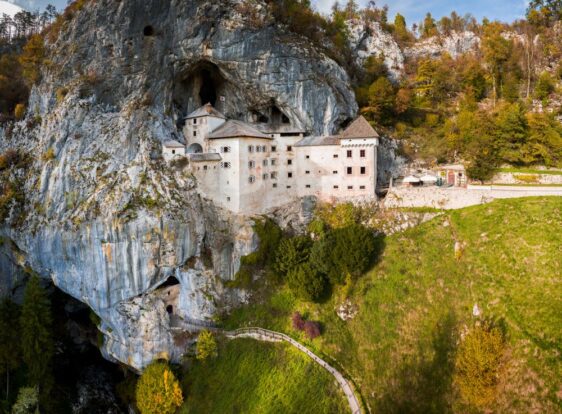 V visoko kamnito steno vklesan grad. Bel s sivo streho in polknami, na fasadi pa grb zadnje živeče družine v Gradu. Pod njim pa se razteza pot v Jamo pod Predjamskim gradom.