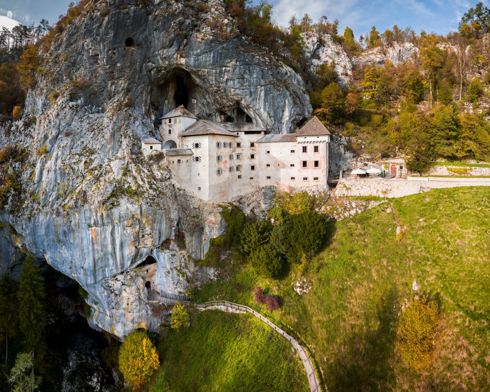 Predjama Castle is built into a towering rock face, its white façade, grey roof, and traditional shutters blending seamlessly with the cliff. The coat of arms of the castle’s last noble family adorns the façade, while below it lies the entrance to the Cave beneath Predjama Castle.