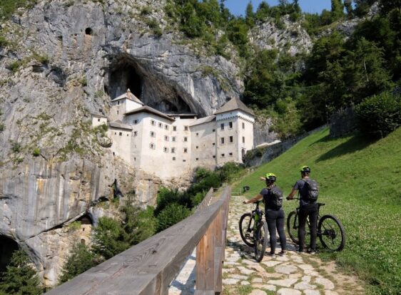 Two cyclists standing at a viewing platform overlooking Predjama Castle, built into a rocky cliff above a cave.