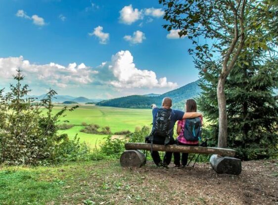 Two hikers sit on a wooden bench at the edge of a forest, looking out across a vast green meadow. In the background, rolling hills and forests rise beneath a blue sky dotted with clouds.