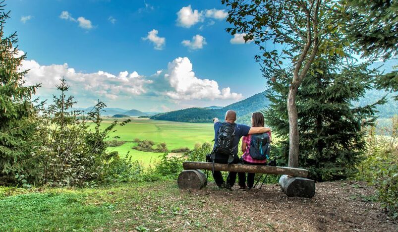 Two hikers sit on a wooden bench at the edge of a forest, looking out across a vast green meadow. In the background, rolling hills and forests rise beneath a blue sky dotted with clouds.