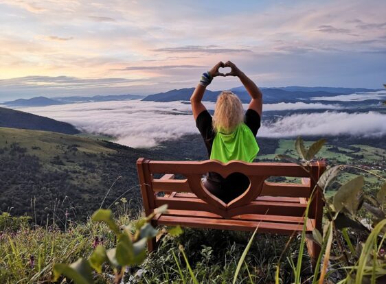 A person sits on a wooden bench at St. Lawrence, forming a heart shape with their hands towards the view. Below lies a mist-covered valley, with rolling hills rising in the distance.