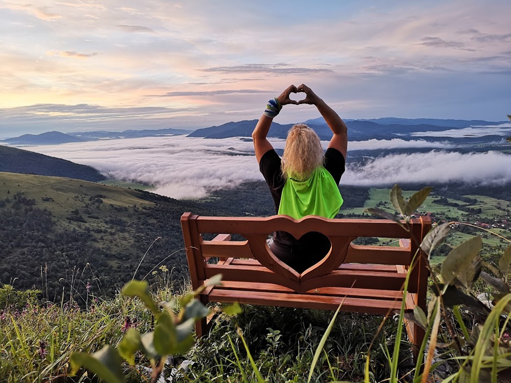 Na fotografiji oseba sedi na leseni klopi na Lovrencu in z rokami oblikuje srce proti razgledu. Pod njo se razprostira dolina, prekrita z meglicami, v ozadju pa se dvigajo hribi.