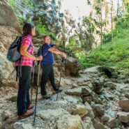 Two hikers walk along a rocky path in Rakov Škocjan. They are surrounded by steep rock walls and forest, while the dry streambed below is scattered with boulders.