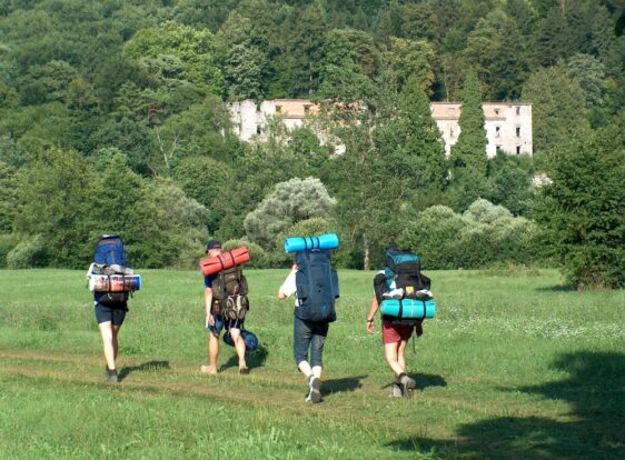 Hikers carrying large backpacks walk along a farm track across the open landscape of Planina Polje.