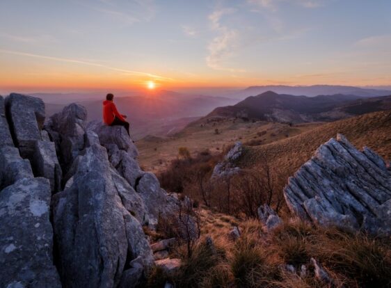A hiker stands on a rocky viewpoint along the trail to Nanos from Razdrto, watching the sun set over the expansive Karst landscape.