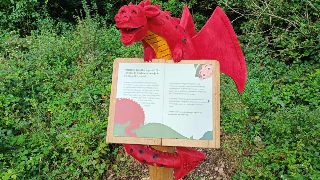 A wooden sculpture of a red dragon stands beside an interpretive trail near Postojna, accompanied by an information panel telling the story of Shepherd Jakob.