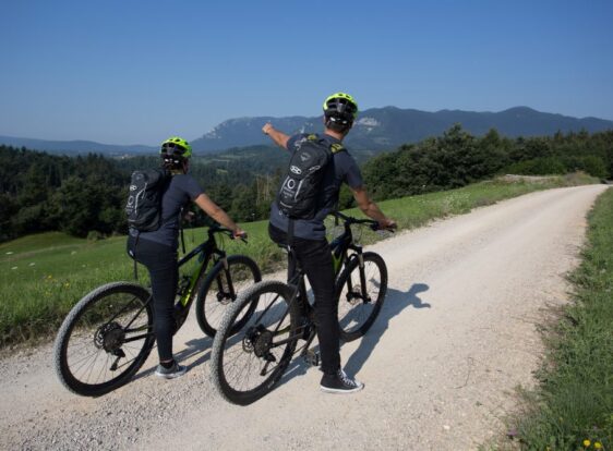 Two cyclists standing beside their bicycles on a gravel road, with one pointing towards the distant hills.