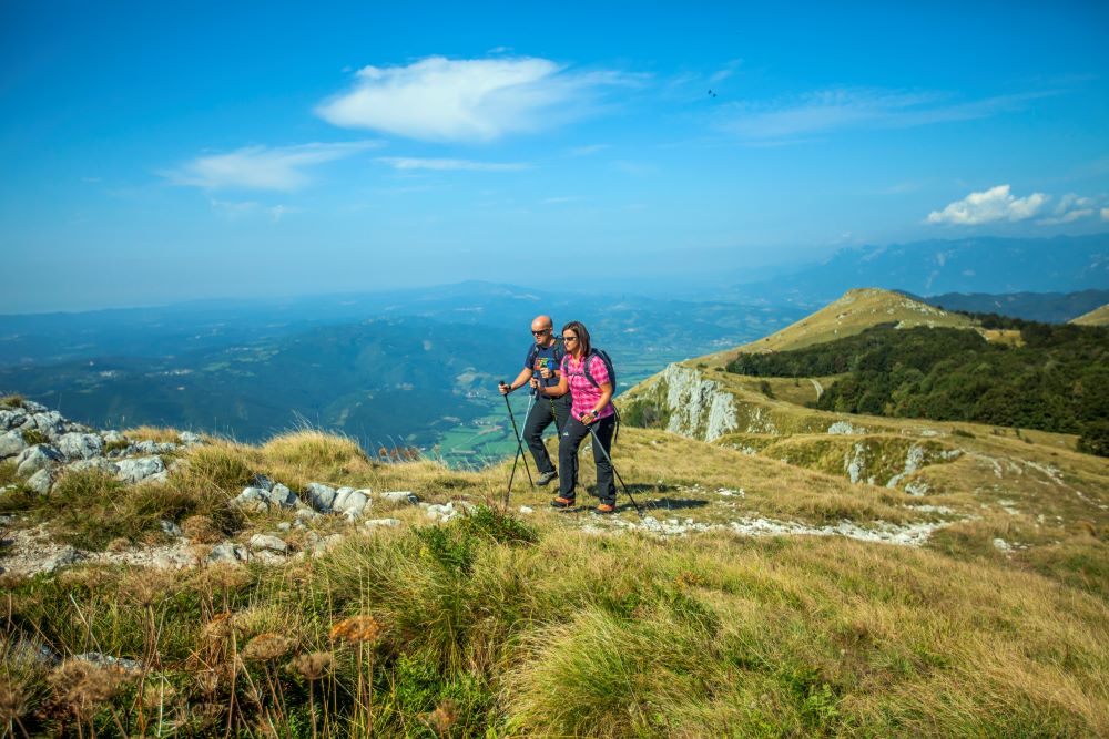 Two hikers walk along the ridge of Nanos, overlooking the valley and the surrounding mountain peaks.