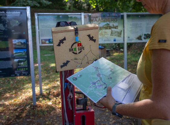 A woman wearing a yellow T-shirt is holding a map beside a bicycle repair station and a secure lockbox. Informational signs are visible in the background.