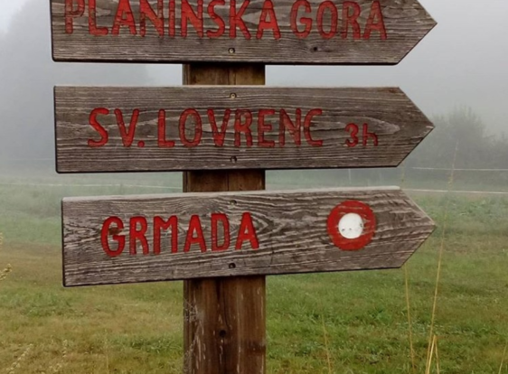 A wooden trail sign stands in a misty meadow, pointing towards Planinska Gora, St. Lovrenc, and Grmada.