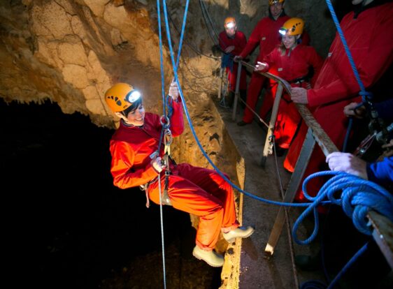 Jamski treking v Postojnski jami. Skupina jamskih navdušencev se s pomočjo vrvi in izkušenih jamarjev spuščajo v podzemlje.