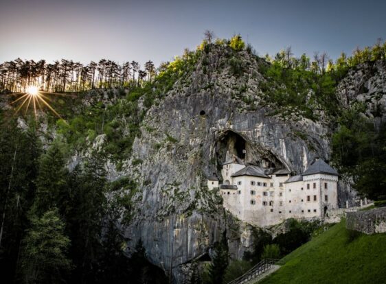 The photograph shows Predjama Castle at sunset, dramatically emerging from the towering cliff face.