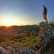 Na fotografiji pohodnik stoji na skalnem razgledišču na Soviču nad Postojno in opazuje sončni zahod. Spodaj se razprostira mesto, okoli pa gričevnata, gozdnata pokrajina.
