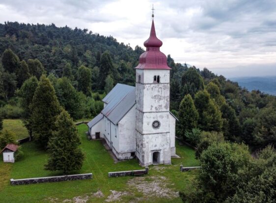 Bela cerkev na Planinski gori, z rdečim zvonikom. Obdana s kapelico in zeleno okolico.
