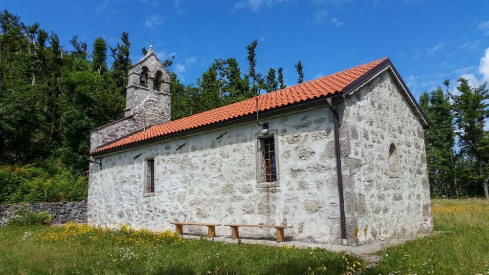 A small stone church with a red roof stands atop St Lovrenc Hill, surrounded by open meadows.