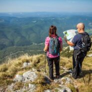 Two hikers stand on the scenic ridge of Mount Nanos, facing the valley as they study a map. Around them are dry karst grasslands and rocky terrain, while a vast forested landscape stretches out in the background.