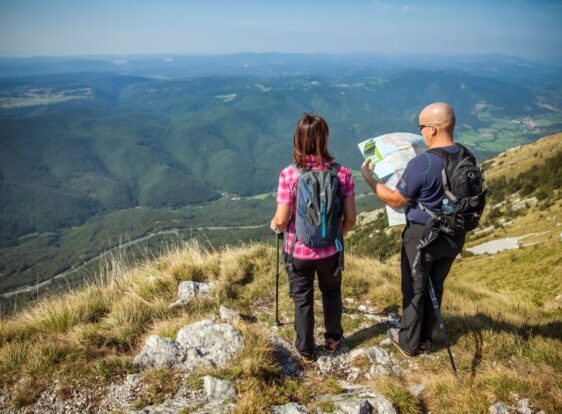Two hikers stand on the scenic ridge of Mount Nanos, facing the valley as they study a map. Around them are dry karst grasslands and rocky terrain, while a vast forested landscape stretches out in the background.
