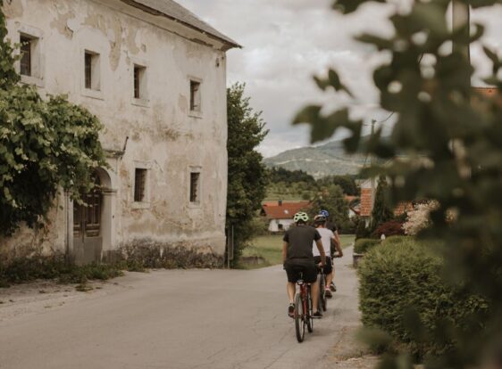 Three cyclists are riding along a paved road past the historic toll house in Landol. An old stone building covered with climbing vines stands beside the road.