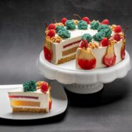 Okrogla torta na belem podstavku, z enim odrezanim kosom na ločenem krožniku. Vidne so več plasti – na dnu svetlo rjav sloj, nad njim svetlejša plast, tanek rdeč sloj in na vrhu gladka svetla krema. Torta je okrašena z majhnimi zelenimi kupčki, koščki sadja in oreščki, ob robu pa visijo tanke rezine sadja.