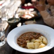 The photograph shows a plate of traditional potato dumplings with a rich meat sauce served on a wooden table at Mladika hut. Glasses and a water carafe are placed beside the meal, while visitors are enjoying their food in the softly blurred background. The image captures the warm and inviting atmosphere of dining on the lodge's terrace.