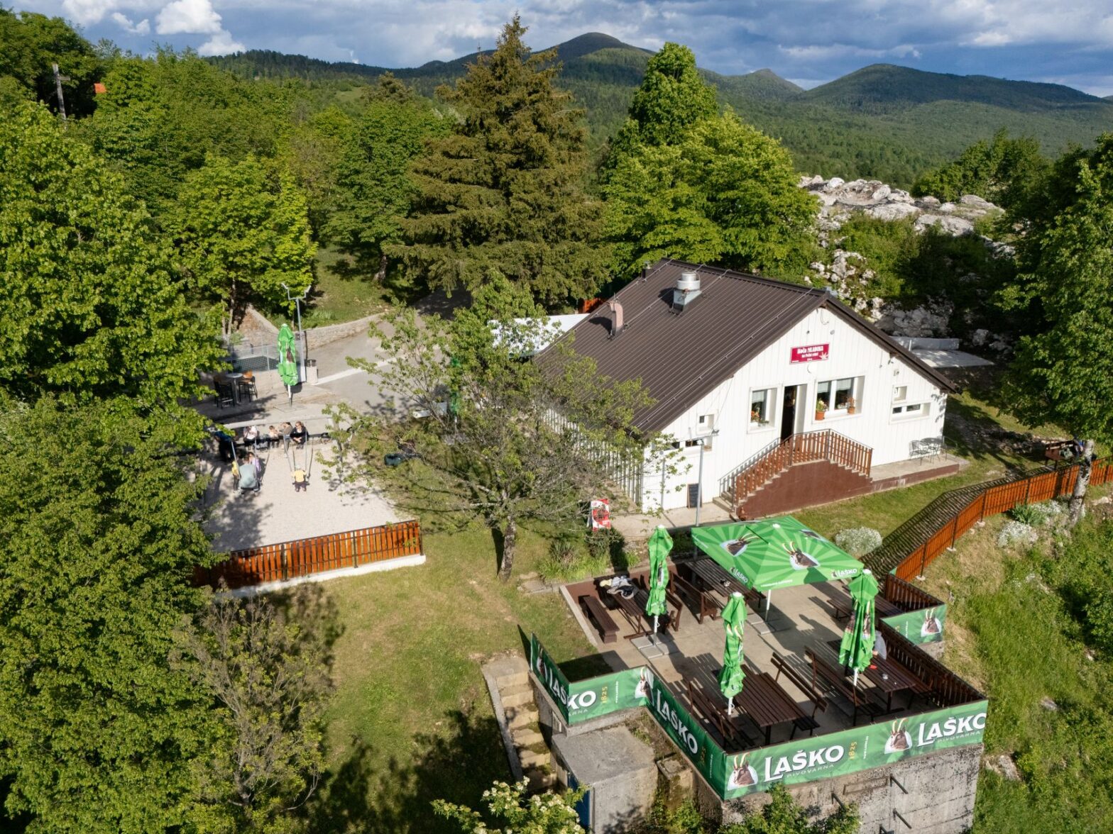 Aerial view of Koča Mladika near Planina, surrounded by the green forests and rolling hills of the Postojnsko destination, with its outdoor terrace and unspoilt nature in the background.