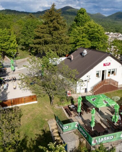 Pogled iz zraka na Kočo Mladika pri Planini, obdano z zelenimi gozdovi in hribi Postojnskega, z zunanjo teraso in naravo v ozadju.