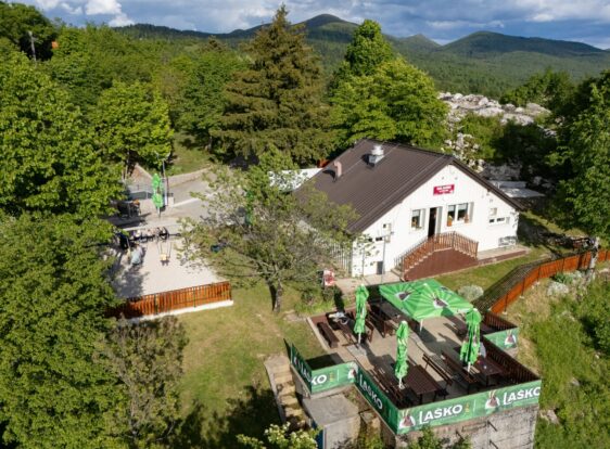 Aerial view of Koča Mladika near Planina, surrounded by the green forests and rolling hills of the Postojnsko destination, with its outdoor terrace and unspoilt nature in the background.