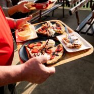 The photograph shows several trays of freshly prepared desserts being served on the terrace of Mladika Hut. The plates are topped with strawberries, whipped cream and chocolate sauce, while visitors are seated at wooden tables in the background. The sunny outdoor setting creates a warm and welcoming atmosphere.