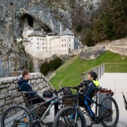 The photograph shows two cyclists seated at a table with their mountain bikes, enjoying the spectacular view of Predjama Castle, dramatically built into the towering limestone cliffs of the Karst landscape.