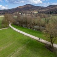 Aerial view of cyclists riding along a gravel road through a landscape of meadows and trees, with the ruins of a historic manor house visible in the background.
