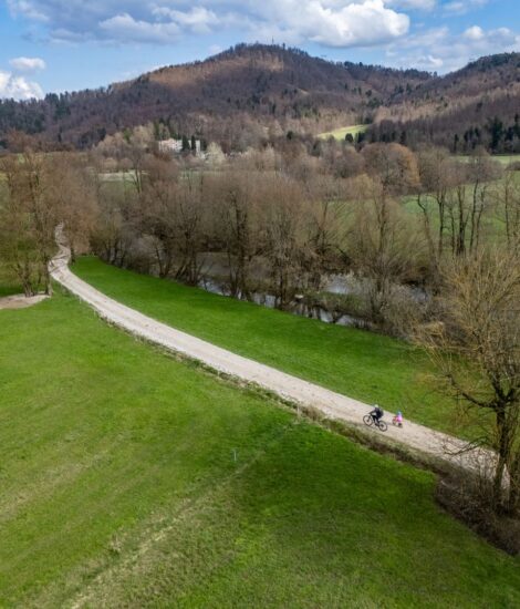 Kolesarji na makadamski poti, ob poteh so travniki in drevesa, v ozadju ruševine dvorca. Fotografija je slikana iz zraka.
