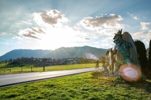 The photograph shows a quiet country road winding through an open rural landscape, with two cyclists riding side by side. On the right, a large sculpture of the Postojna Cave dragon stands on a grassy verge beside the road, accompanied by the shepherd boy Jakob. Meadows, traditional houses, and rolling hills stretch across the background beneath a bright sky with scattered clouds, while the low sun casts a warm glow over the landscape.