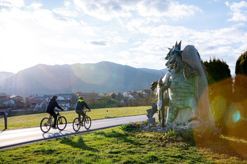 The photograph shows two cyclists riding side by side along a quiet country road through an open landscape. On the right, a large sculpture of the Postojna Cave dragon stands in a meadow beside the road, accompanied by the shepherd boy Jakob. Meadows, traditional houses, and rolling hills stretch across the background beneath a blue sky with scattered clouds, while the low sun shines towards the camera, bathing the scene in warm light.