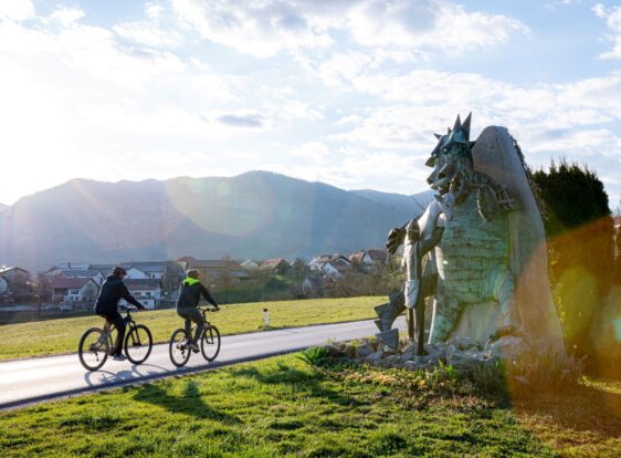 The photograph shows two cyclists riding side by side along a quiet country road through an open landscape. On the right, a large sculpture of the Postojna Cave dragon stands in a meadow beside the road, accompanied by the shepherd boy Jakob. Meadows, traditional houses, and rolling hills stretch across the background beneath a blue sky with scattered clouds, while the low sun shines towards the camera, bathing the scene in warm light.