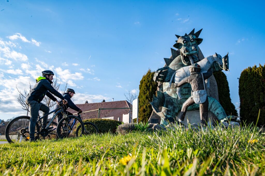 The photograph shows two cyclists standing beside their bicycles in a meadow, admiring a sculpture. The sculpture depicts the dragon of Postojna Cave with its mouth wide open, accompanied by the shepherd boy Jakob carrying his satchel. Traditional houses, trees, and a blue sky with scattered clouds form the backdrop.