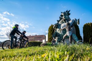 The photograph shows two cyclists standing beside their bicycles in a meadow, admiring a sculpture. The sculpture depicts the dragon of Postojna Cave with its mouth wide open, accompanied by the shepherd boy Jakob carrying his satchel. Traditional houses, trees, and a blue sky with scattered clouds form the backdrop.